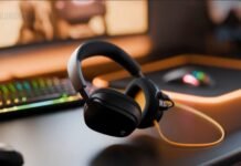 Over-ear black gaming headphones resting on a desk with a neon RGB keyboard blurred in the background and a yellow audio cable.