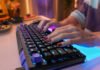 Close-up of hands typing on a backlit mechanical keyboard with purple and blue LEDs against a blurred colorful desk.