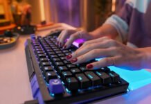 Close-up of hands typing on a backlit mechanical keyboard with purple and blue LEDs against a blurred colorful desk.