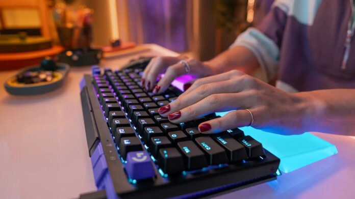 Close-up of hands typing on a backlit mechanical keyboard with purple and blue LEDs against a blurred colorful desk.