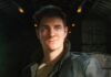 Young man with a backpack gazing upward in a dark, industrial-looking hangar or warehouse setting.