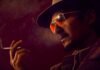 Close-up of a man wearing a hat and sunglasses, smoking a cigarette in a moody red-lit scene with visible smoke.