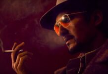 Close-up of a man wearing a hat and sunglasses, smoking a cigarette in a moody red-lit scene with visible smoke.