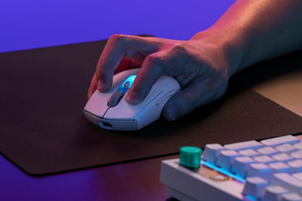 Close-up of a hand resting on a white wireless mouse with a blue LED, on a dark desk with a keyboard in the foreground under colorful lighting. G305 X Superlight LIFE White Alt