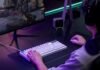 Gamer wearing a headset using a white keyboard and mouse, playing a shooter on a large monitor with neon purple lighting.
