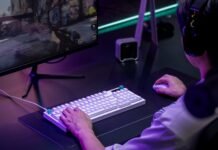 Gamer wearing a headset using a white keyboard and mouse, playing a shooter on a large monitor with neon purple lighting.