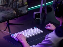 Gamer wearing a headset using a white keyboard and mouse, playing a shooter on a large monitor with neon purple lighting.