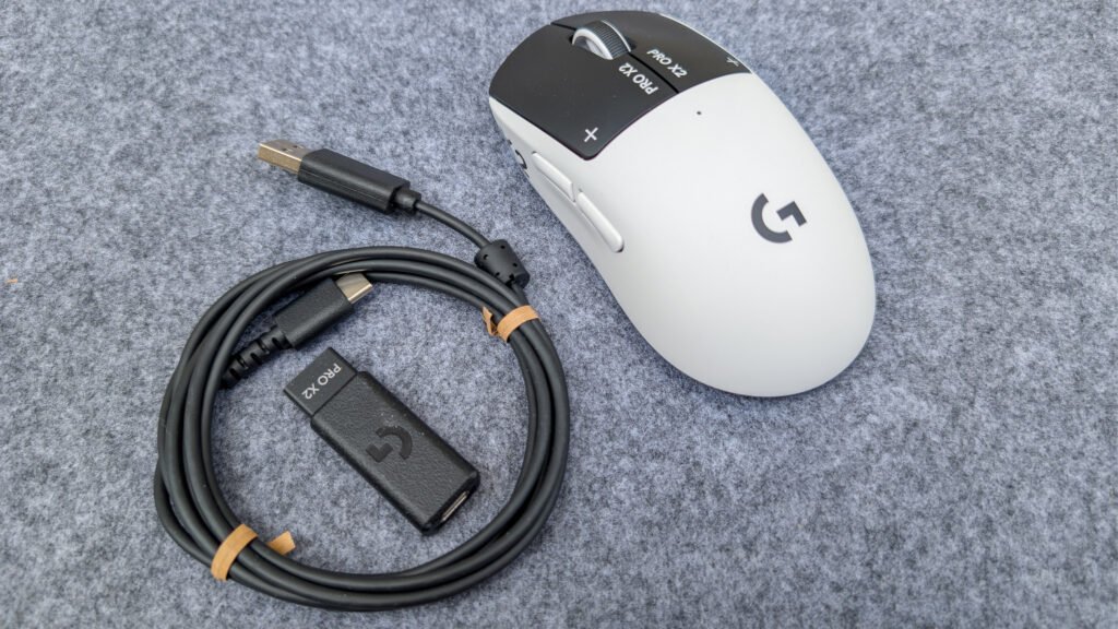 White Logitech Pro X gaming mouse with a long USB-C cable laid on a gray textured surface, showing the logo and side buttons. PXL_20260307_104333513RAW-01COVER