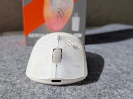 White wireless computer mouse on a gray carpet with a blurred orange and gray product box in the background.