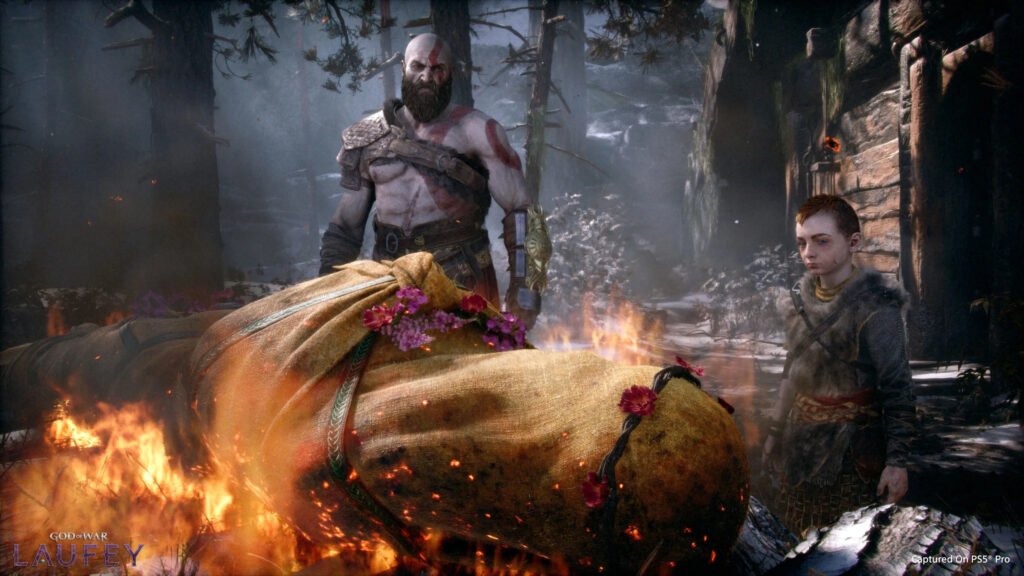 god of war laufey 2 - GamersPack ישראל Kratos, with red tattoo and beard, stands beside a large flower-adorned sack in a snowy forest ruin, while a young boy looks on amid flames and smoke. god of war laufey 2