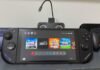 Nintendo Switch OLED in handheld mode on a desk, displaying the home screen with game icons and two controllers on either side.