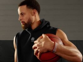Athlete in a black sleeveless hoodie holding a basketball against a gym background, looking to the left.