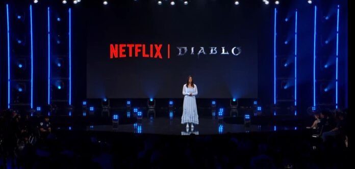 Blizzcon 2026: דיאבלו תשתף פעולה עם נטפליקס בסדרת אנימציה חדשה Presenter stands on stage with a large screen showing ‘NETFLIX | DIABLO’ behind her at a reveal event. Audience in foreground, blue stage lighting.