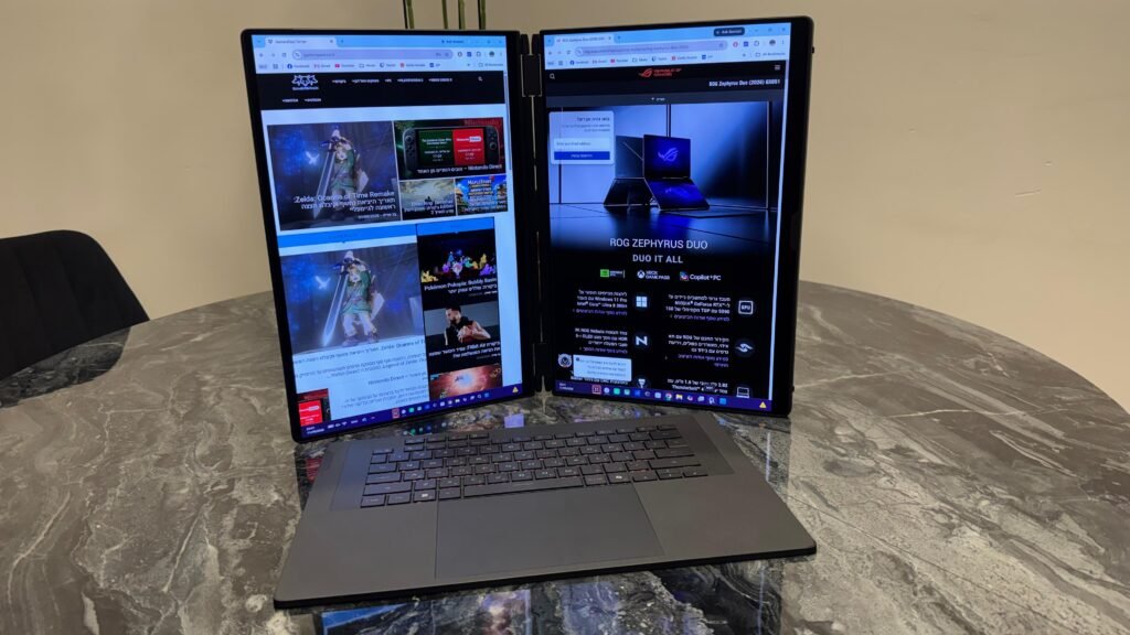Dual-screen foldable laptop in tent mode on a marble table, showing two browser windows with Hebrew text and imagery. WhatsApp Image 2026-09-22 at 112537 1