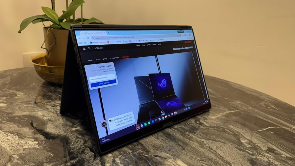 Foldable ASUS 2-in-1 laptop on a marble table, screen showing a tech site with Hebrew text, plant in the background. WhatsApp Image 2026-09-22 at 112537 3