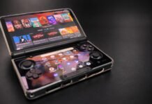 Foldable handheld gaming console open on a dark surface, showing colorful game icons on the top screen and control buttons below.