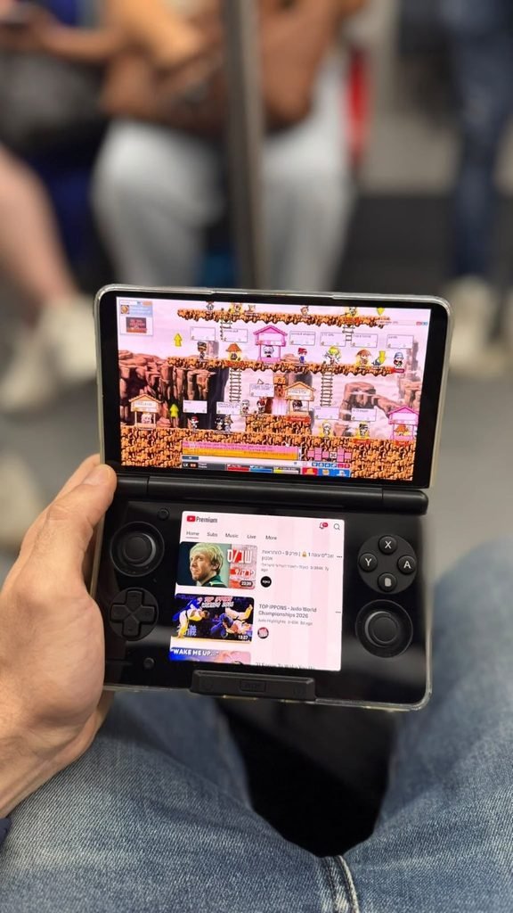 fc34921a-99a1-4655-9716-b9bb1055980d - GamersPack ישראל Person holds a dual-screen handheld console showing a colorful pixel-art game on the top screen and a video app on the bottom screen. fc34921a-99a1-4655-9716-b9bb1055980d